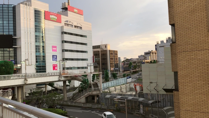 左に白いビルが建ち、右に茶色のビルが建つ写真。ビルの間を通る白い歩道橋がある。