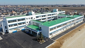 白いコンクリートでできた外壁と緑色の屋根をもつ大きな学校が広いグラウンドにある