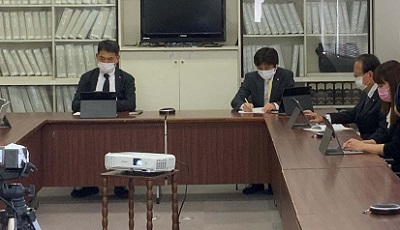 黒いスーツを着た男性3人とスーツを着てピンクのマスクをつけた長髪の女性が椅子に座り勉強会を行っている