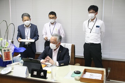議長室にて、黒いスーツを着た短髪の男性が着席し、その後ろに短髪の男性3人が立って、机に置いてあるタブレットを見ている