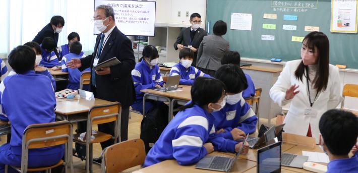 中学校の教室で、立っている男性と女性の議員が、生徒の近くで話しかけている