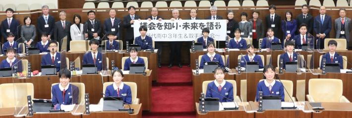 市議会の議場で、模擬議会に参加した中学生が議席に座り、その後ろに議員が並んでいる