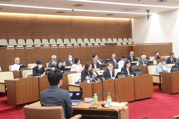 議場の座席に21名の男性と女性の議員が座って話を聞いている