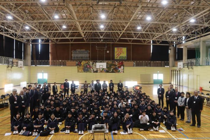 中学校の体育館に議員と生徒、数十名が並んでいる