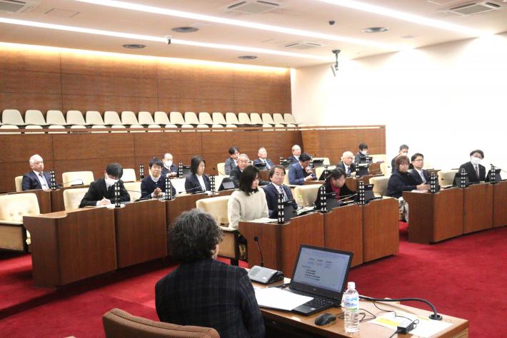 議場の議席に十数名の議員が座り、座っている男性の講師の話を聞いている