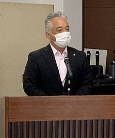 大会議室に茶色い台が置かれ、スーツを着た短髪の男性が立っている