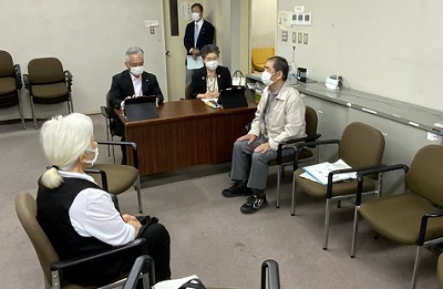 会議室内に椅子が置かれ、スーツを着た短髪の男性と女性が1人ずつ、その手前に私服姿の男性と女性が1人ずつ、計4人が座っっている