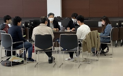 茶色い壁を背景に、椅子が円形に置かれ、5人の男性と1人の女性が向かい合って座っている