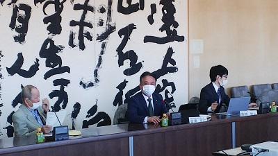 横一列の机に3人の男性が座っている。中央の男性議員(金澤委員長)が着座のまま挨拶をしている写真