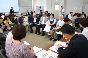 会議室にて、たくさんの男女が紙の資料を持って、輪になって座って話している