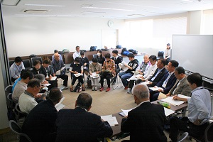 会議室の中央に、たくさんの男女が輪になって座り、手元の紙の資料を見ながら話している
