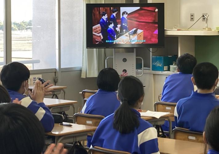 教室で生徒が席に座って、前方のモニターに映る映像を見ている