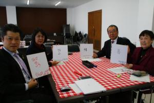 漢字1文字が書かれた色紙を手に持ってカメラに目を向ける議員4人（グループ2）の写真。