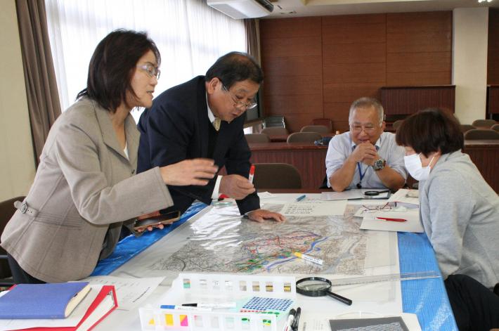 男性議員2人(関戸・小堤議員)、女性議員2人(池田・加増議員)が机の中央に置かれた地図を囲んで話し合いをしている