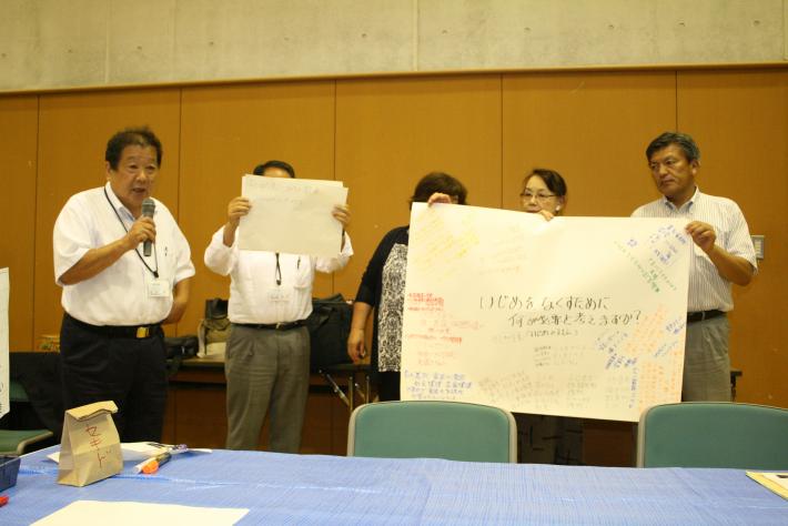 グループ「答弁」の発表の様子。意見をまとめた模造紙と紙を手にする参加者とマイクを手にした発表者が起立して発表している。