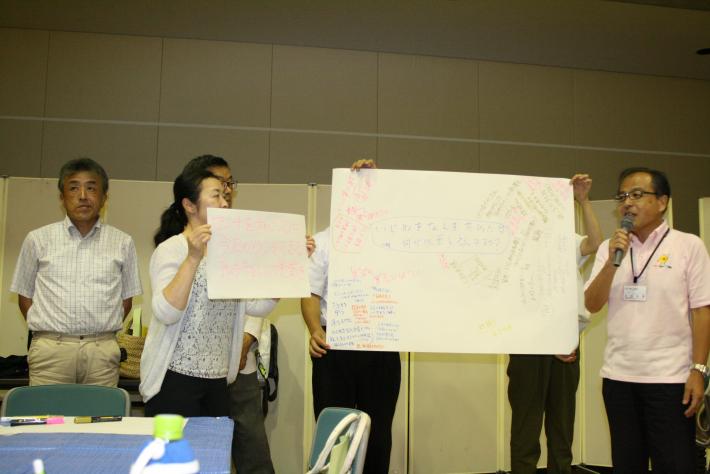 グループ「地域密着」の発表の様子。意見を記載した模造紙を手に掲げる横で発表者が話をしている。