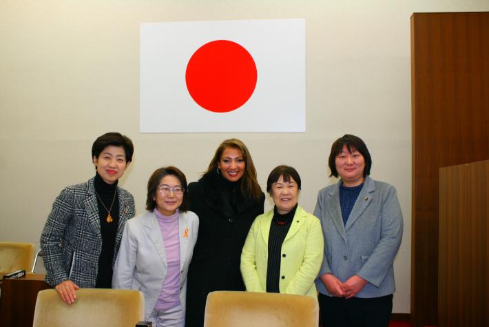 日本の国旗が飾られた壁を背景に、長髪の女性1人と短髪の女性4人が笑顔で立っている