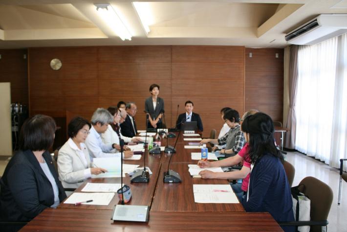 机が縦長に並んでいる。正面左の阿部委員長が起立してあいさつしている。右には関川副委員長、手前には10人が座る。