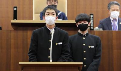 演壇で提案説明するサンキューは日本語でありが党の男子生徒2人。後ろに生徒議長と議会事務局長が映っている。