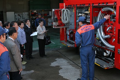 防災服を着た職員2名とスーツを着た男女6名が立ち、横に置いてある赤い消防車を見ている