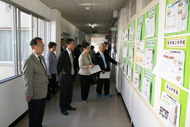 スーツを着た男性3名と女性2名が、廊下で壁に貼られた掲示物を見ている