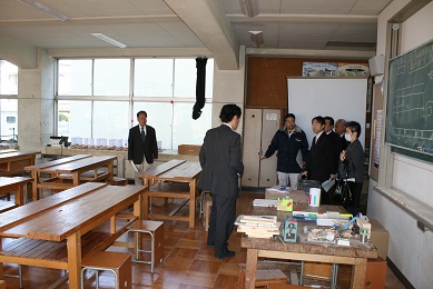 学校の教室内に、スーツを着た男性5人と女性1人、ジャンパーを着た男性1人が立っている。教室には木製の縦長の机が6つ置かれている