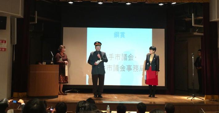 舞台の上に、学生服を着た男性1名とスーツを着た女性と男性1名ずつが立っている