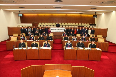 議場の議席に制服姿の学生20名が座り、その奥に20名のスーツ姿の男女が立っている。傍聴席に1名の男性が座っている