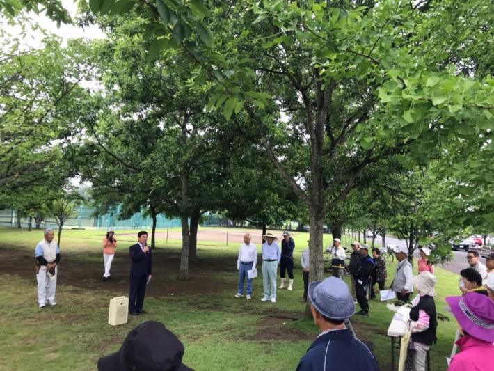 木々が並ぶ広場に参加者が集まっている。その前で市長があいさつをしている様子。