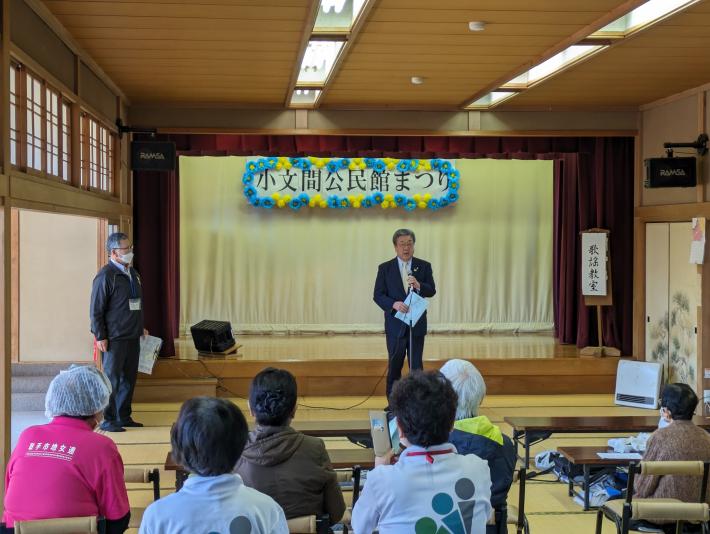 和室ステージ前にて着座の参加者へ挨拶をする市長。背景には「小文間公民館まつり」