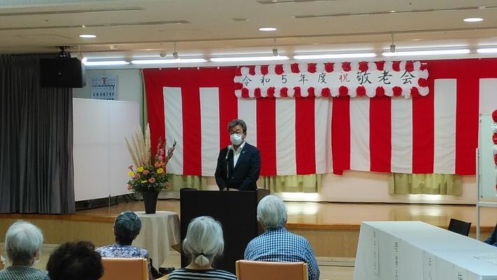 紅白垂れ幕を背にマイクで挨拶をする市長。垂れ幕上に「令和5年度祝敬老会」の横断幕