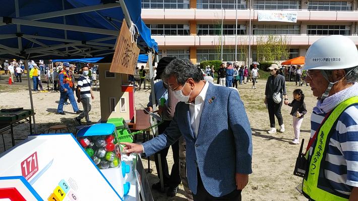 校庭でテント下に置かれたカプセルトイに手を伸ばす市長