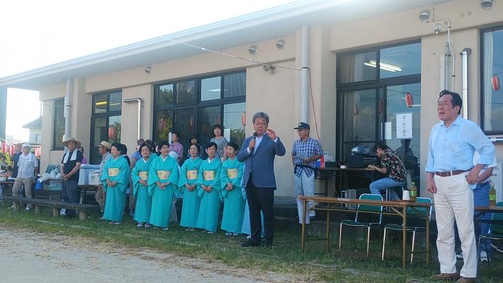 相馬南公民館夏まつり盆踊り大会での挨拶の様子。建物前にそろいの浴衣をきたひと立ちが並び、その横でマイクを持つ男性が挨拶している。