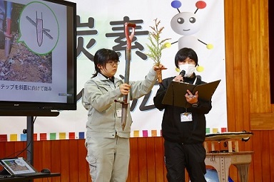 県南農林事務所の職員が右手に穴掘り器、左手にスギの苗木を持ちながら、スギの植栽方法の説明をしている。