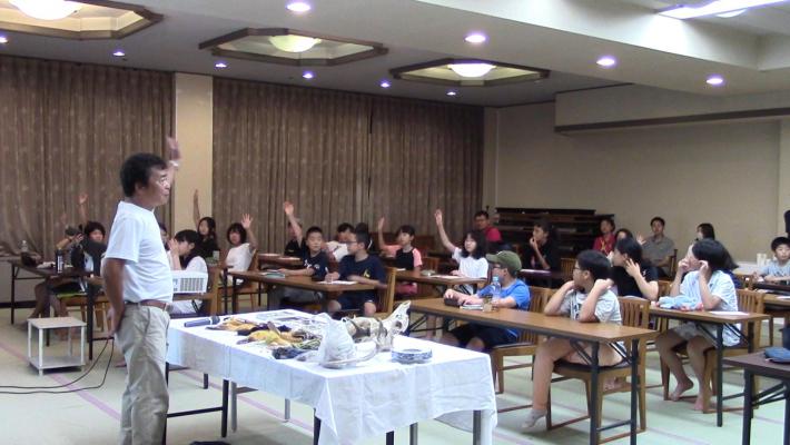 高柳盛芳さんの講話の様子。写真左部分で高柳さんが話しており、写真中央及び右側のこどもたちは高柳さんの質問に手を挙げて答えている。