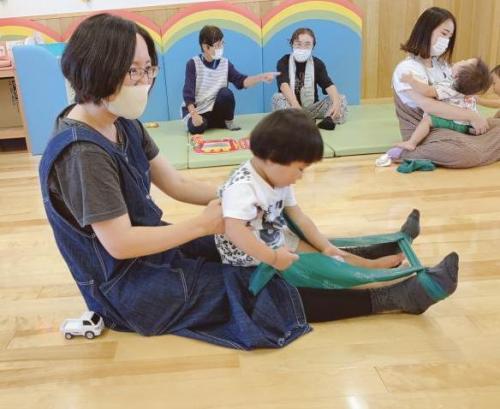 若い女性の膝の上に子どもがすわっている。女性はそのまま脚にひっかけたゴムチューブを伸ばして運動している。