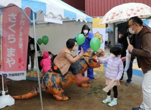 恐竜の着ぐるみを着た女性が小さいお子さんに風船を渡しているところ
