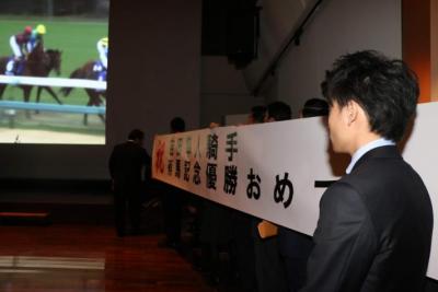 写真右にいる男性が薄暗い中吉田隼人騎手有馬記念優勝おめでとうと書かれた白い横断幕の前で写真左奥で流れる馬が走る映像を見ている画像