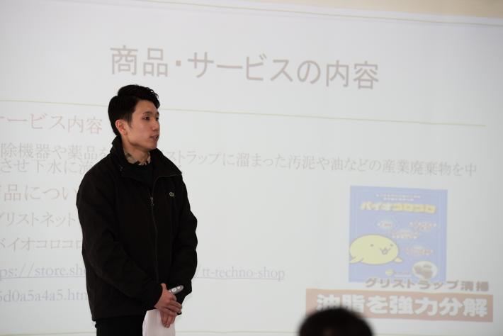 山川拓巳さん発表の様子。黒い上着、短髪の若い男性が、手を体の前に組んで置き、何かを話している。