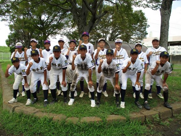 藤代軟式野球クラブの生徒たち。木の前、芝生様の緑の上でユニフォーム姿でガッツポーズをする生徒達。