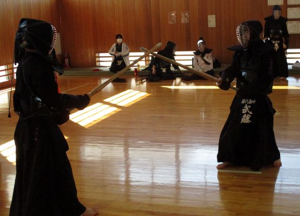 1年生同士の対決のようす。武道場内で対決する2人の剣士と、壁際でそれを見学する面を外した剣士3人の写真。