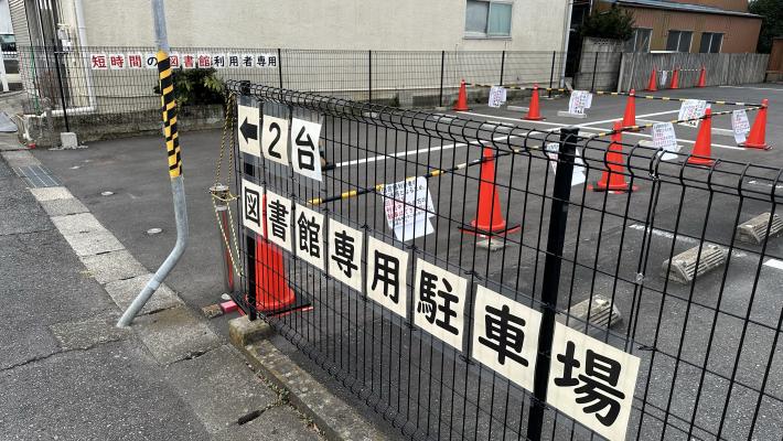 （全体）短時間利用者専用駐車場。フェンスに「←2台図書館専用駐車場」と書かれた紙が貼られた駐車場の写真。