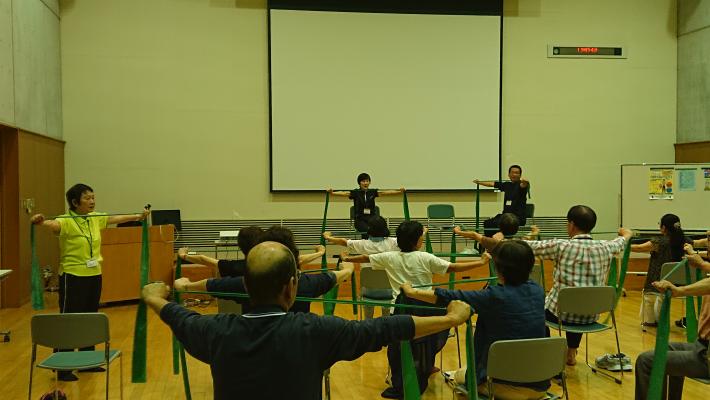 椅子に座って、胸の前でチューブを横に引っ張っている様子