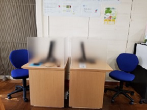 デスクが2台向かってならび、青い事務椅子が設置されている。机上には目隠しのパーティションと端末がある。