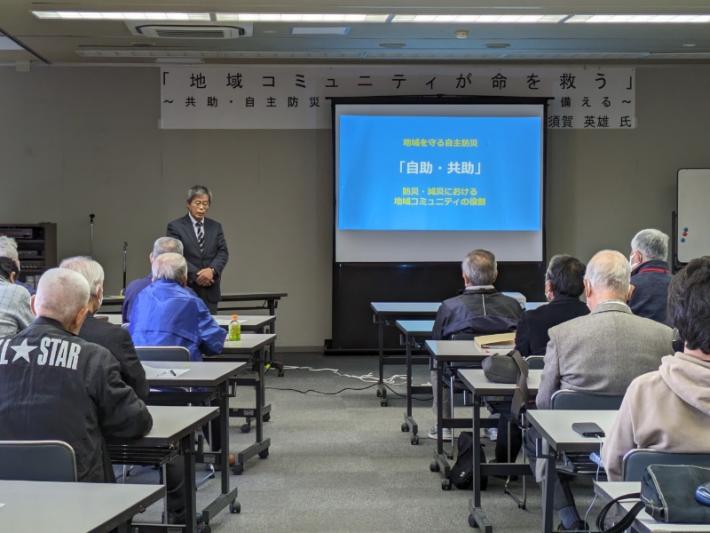 須賀英雄氏プロジェクターを使用した講演写真。プロジェクターに資料を写しだし,講演する講師とそれを着座で聞く市民たち。