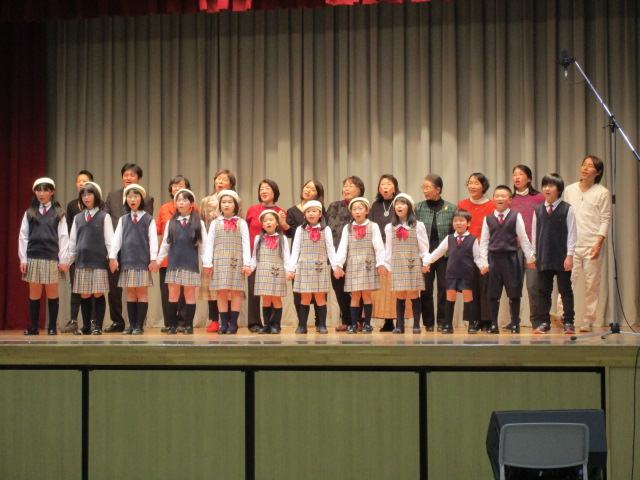 舞台の上で、子ども合唱団12人と大人12人が歌っています。