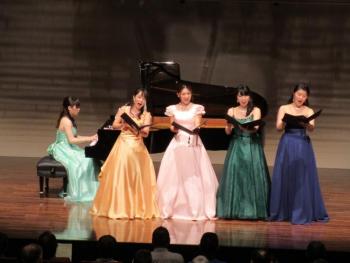 舞台上でピアノ奏者と歌手4人が音楽を披露中。