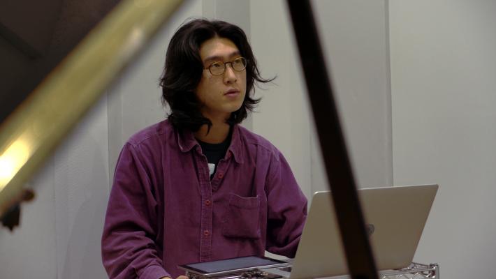 赤いシャツを着た長髪の男性がパソコンを操作し作曲している。藤井登生写真