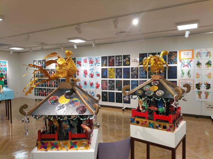 手前に黒い屋根のお神輿が2台、奥の壁にスタンプで作った花の絵の作品が飾られた様子