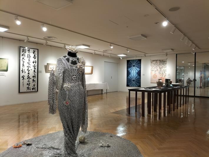 床にはトルソーに着せた工芸作品と展示台があり、奥の壁には書、染め物、織物の作品が展示されている様子